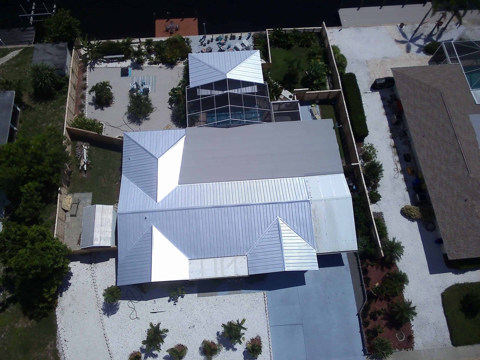 Aerial view of a residential property with a large metal-roofed structure, swimming pool, and landscaped garden. The well-maintained home, a testament to quality roof replacement in Sarasota County, is separated by fences and surrounded by other buildings and green areas.