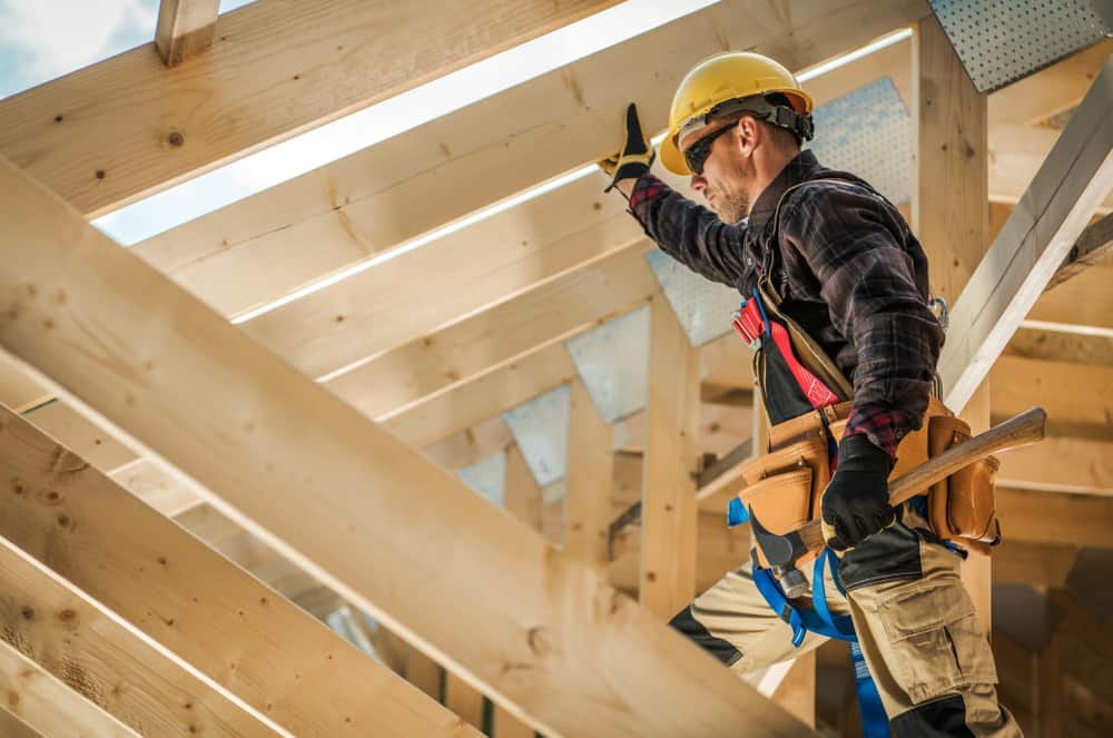 A roofing contractor in Sarasota County, wearing a yellow hard hat, safety harness, and protective gloves, is seen installing wooden beams on a roof. He is raising one arm to secure a beam while holding a hammer in the other hand, surrounded by light-colored wooden trusses.