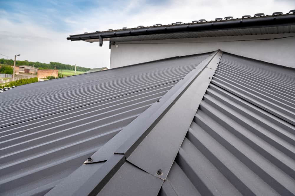 A close-up view of a Metal Roof Sarasota County with ribbed panels and fasteners. The roof has a central ridge cap and is part of a modern building with overhanging eaves and a gutter system. The background includes a partially visible landscape and a cloudy sky.