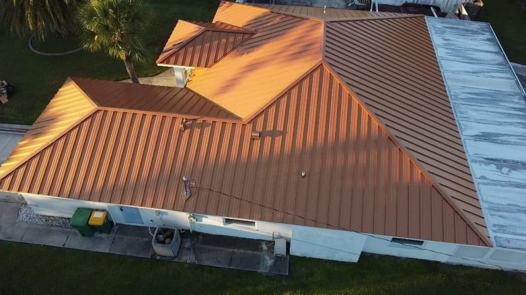Aerial view of a house with a copper-colored metal roof featuring multiple ridges and angles. The house is surrounded by green grass, with a palm tree nearby. A white section of the roof appears older or weathered, suggesting it may need attention from a roofing contractor in Sarasota County.