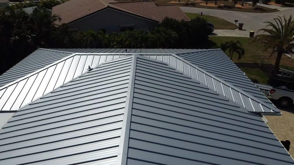 Aerial view of a modern house with a silver metal roof, characterized by clean lines and overlapping panels. The property has surrounding greenery, with additional houses visible in the background. A truck from a roofing contractor in Sarasota County is parked in the driveway on the right side.