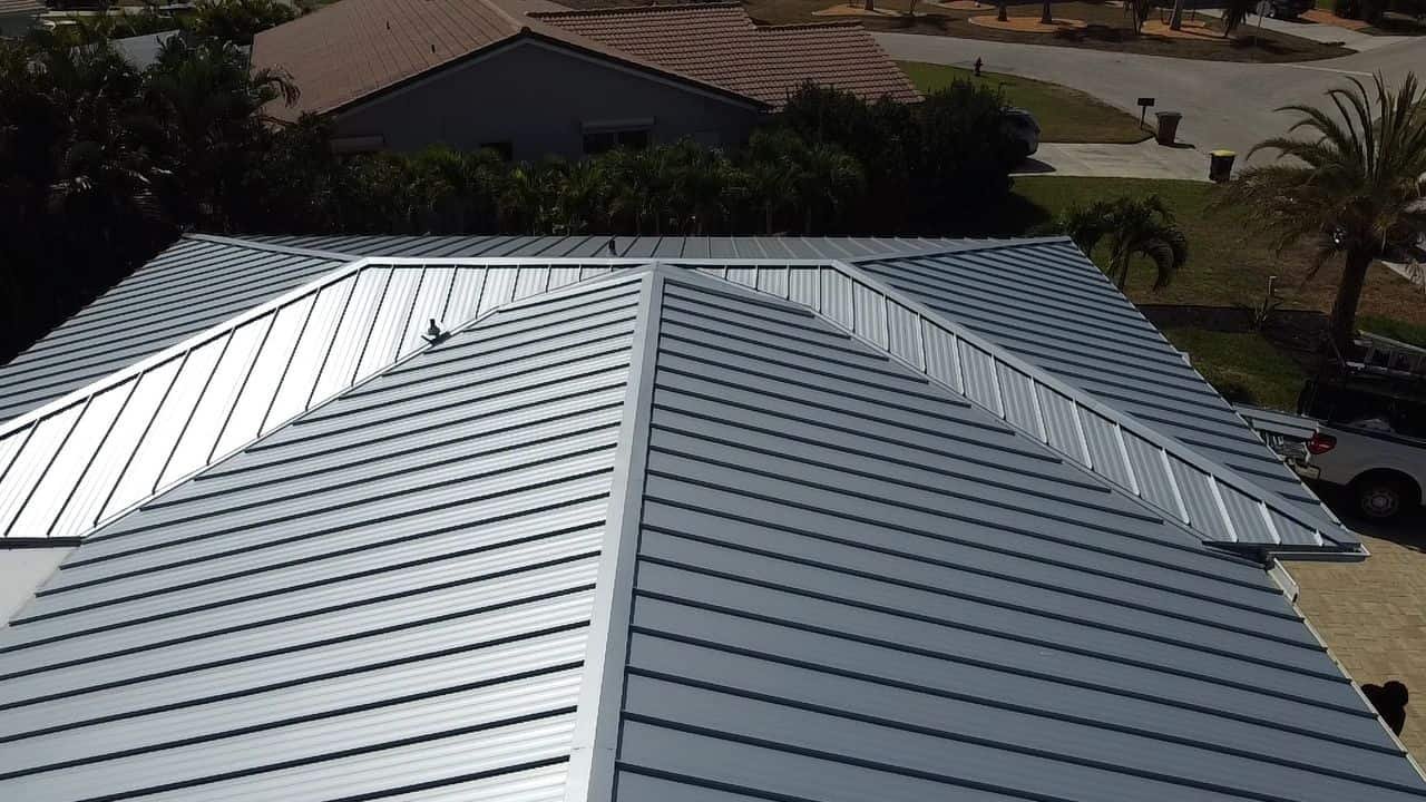 Aerial view of a modern house with a silver metal roof, characterized by clean lines and overlapping panels. The property has surrounding greenery, with additional houses visible in the background. A truck from a roofing contractor in Sarasota County is parked in the driveway on the right side.