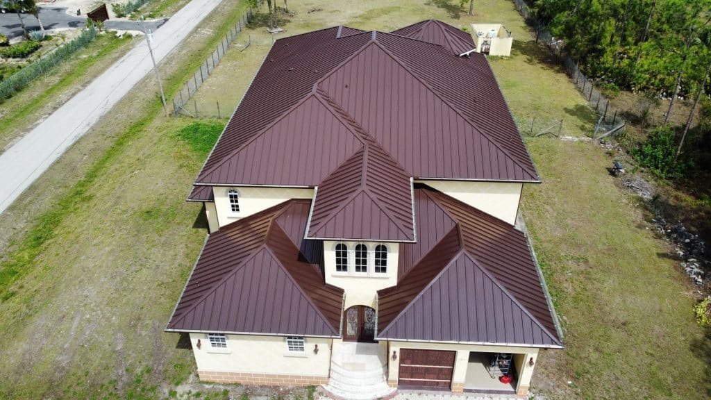 Aerial view of a large, beige, two-story house with a dark brown metal roof completed by a roofer in Sarasota County. The house features multiple gables and dormer windows, with a small front porch and an attached garage. It is surrounded by grass and trees, with a road visible on the left side.