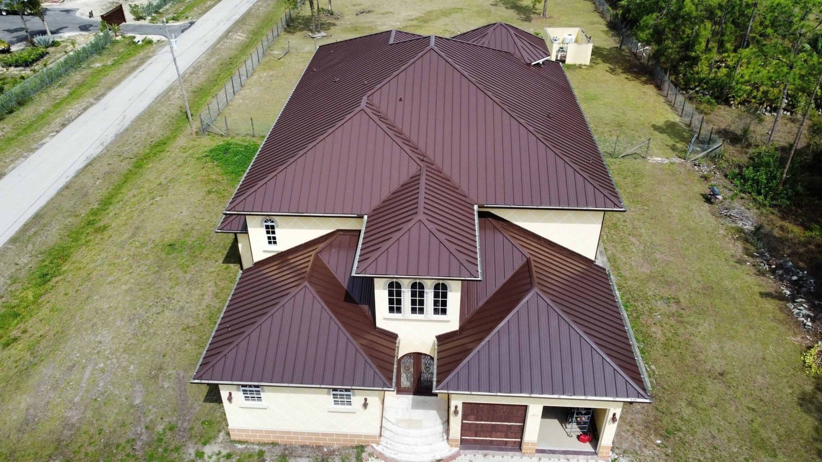 Aerial view of a large, beige, two-story house with a dark brown metal roof completed by a roofer in Sarasota County. The house features multiple gables and dormer windows, with a small front porch and an attached garage. It is surrounded by grass and trees, with a road visible on the left side.