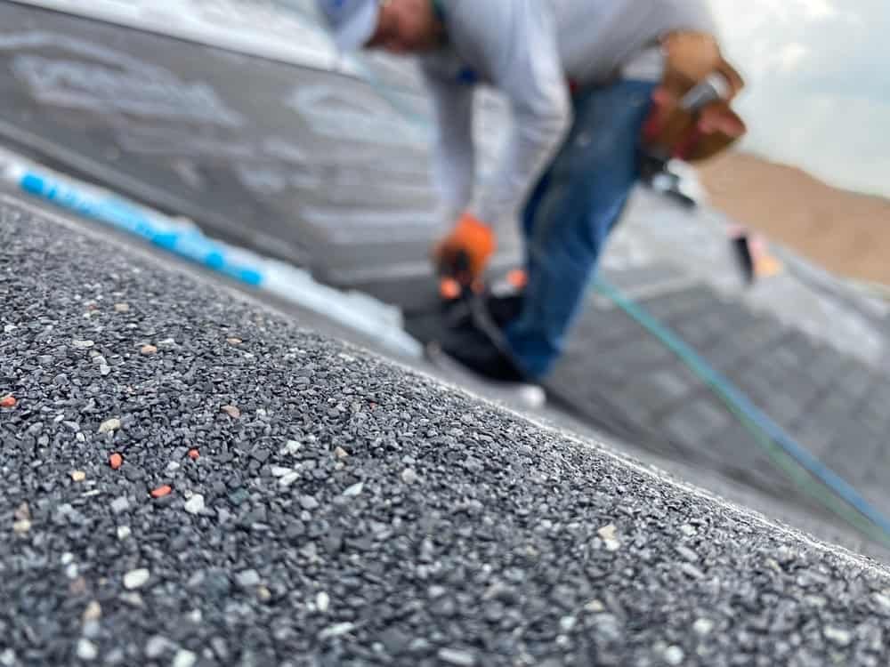 Close-up of a shingled rooftop with a blurred background showing a worker in a white helmet and gray shirt installing roofing materials. The worker is crouched and using tools, with a toolbelt visible at their side.