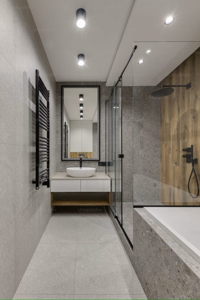Modern bathroom with gray stone tiles, featuring a glass-enclosed shower with black fixtures, a wall-mounted sink with a round white basin, a tall mirror, and a heated towel rack on the left. Recessed ceiling lights provide illumination.