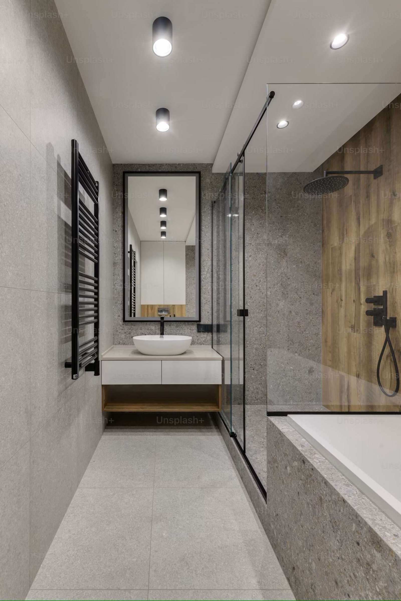 Modern bathroom with gray stone tiles, featuring a glass-enclosed shower with black fixtures, a wall-mounted sink with a round white basin, a tall mirror, and a heated towel rack on the left. Recessed ceiling lights provide illumination.