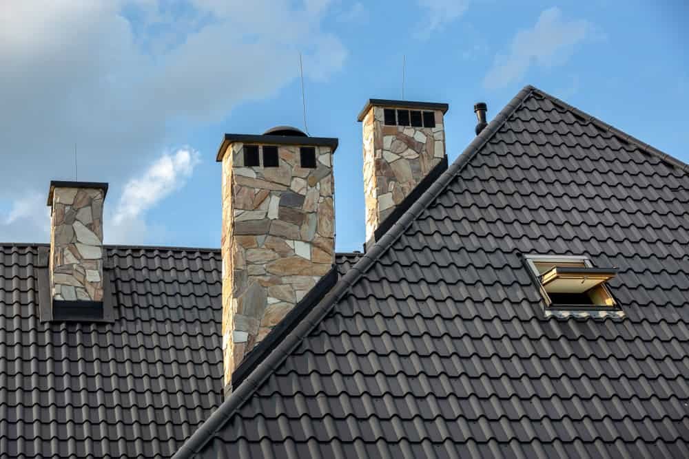 A modern house roof with dark gray, textured tiles features two stone chimneys and a skylight window. The chimneys are crafted from light-colored stones and are evenly spaced. The sky in the background is partly cloudy.