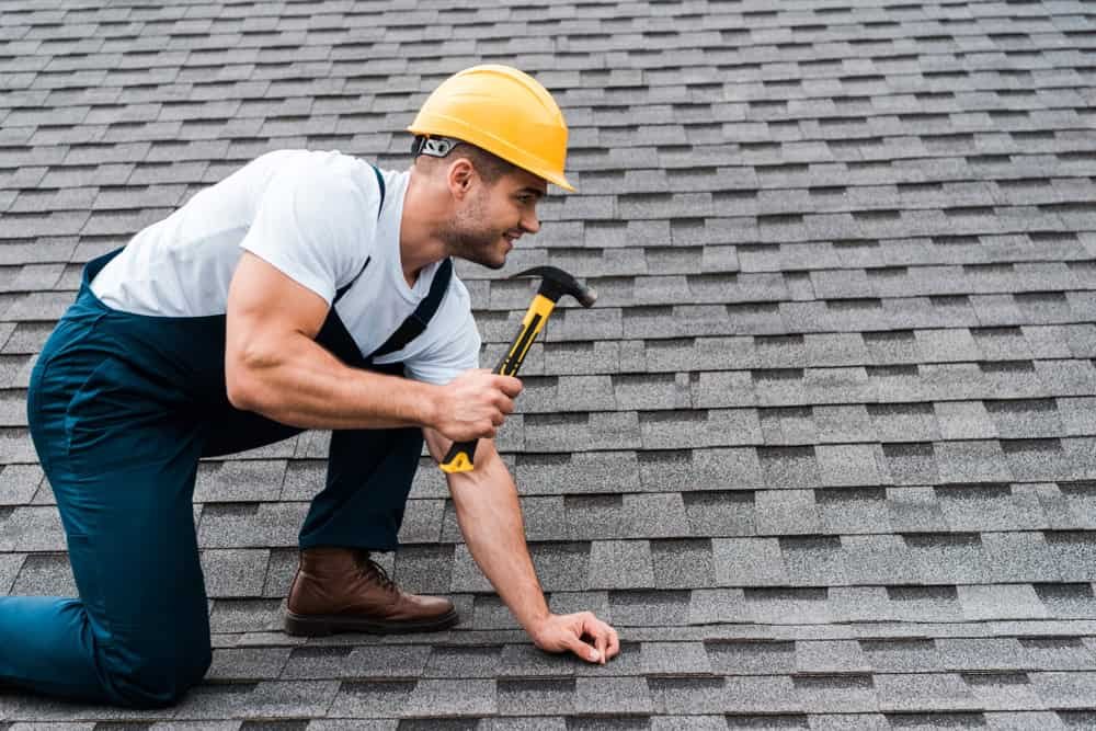 A person wearing a yellow hard hat and blue overalls is kneeling on a shingled roof. They are holding a hammer, appearing to work on the roof. The sky is not visible in the background.