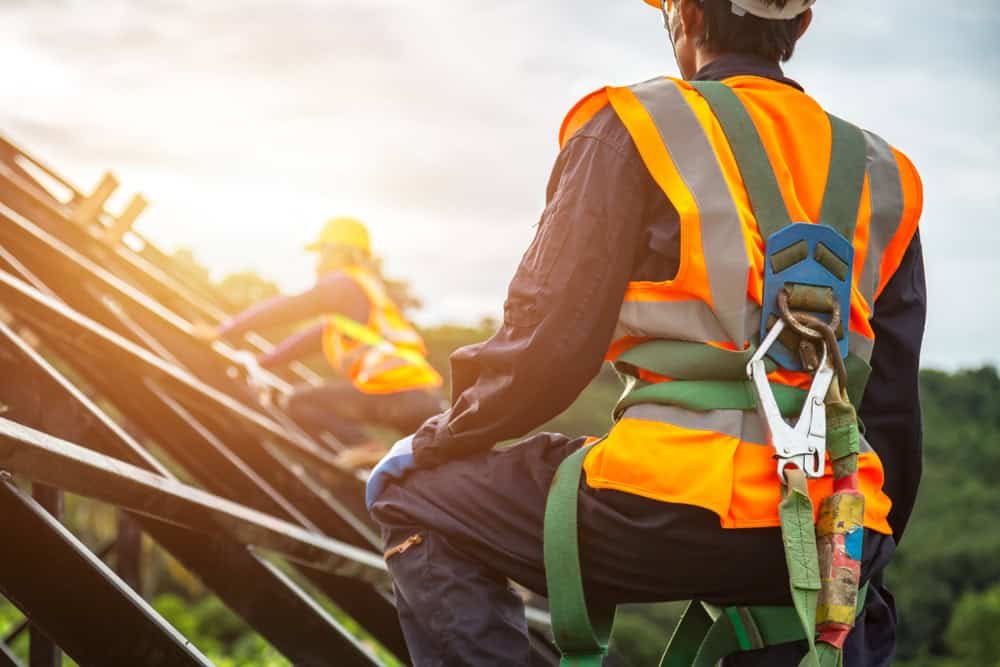 Two construction workers are climbing a metal structure outdoors. They are wearing orange safety vests, helmets, and harnesses. The background features a blurred view of greenery and a bright sky.