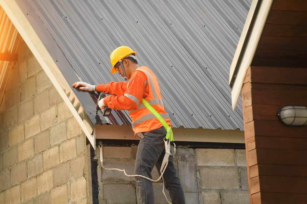 A construction worker wearing an orange safety vest, yellow hard hat, and harness is using a power drill to secure a corrugated metal sheet on a roof. The sun is shining from the top left corner, casting a warm glow.