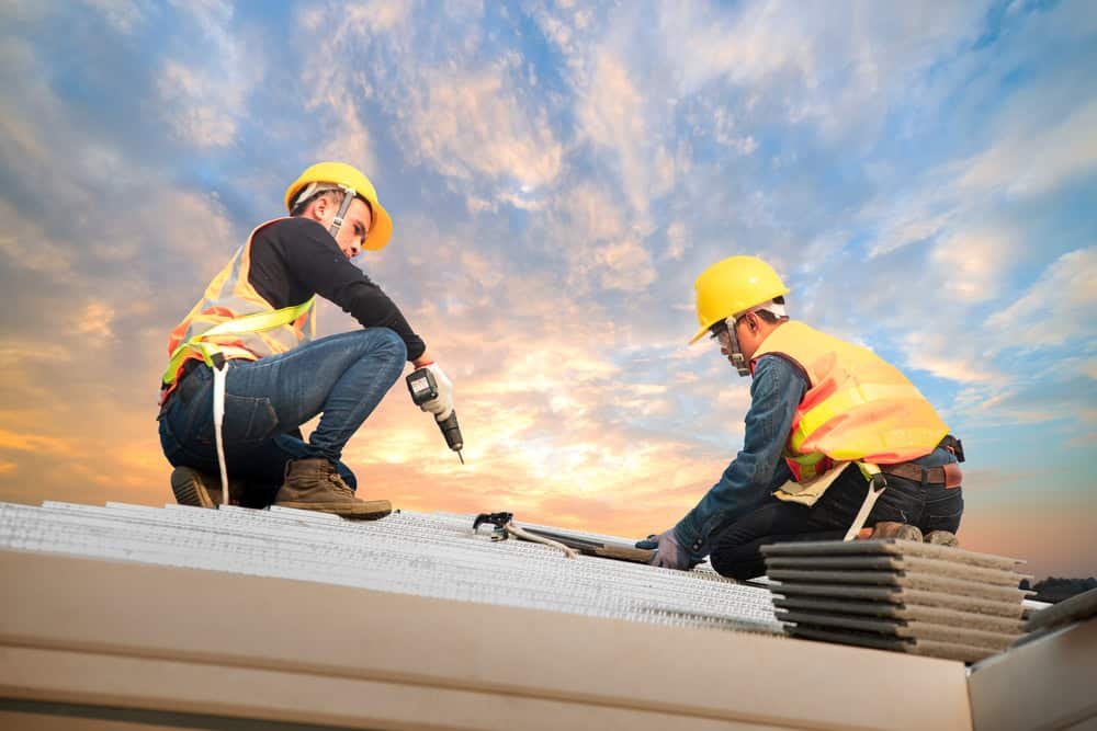 Two construction workers wearing yellow helmets and safety vests are working on a roof at sunset. One is using a power drill, while the other holds a panel. Stacks of roofing materials are nearby, and the sky is partly cloudy.