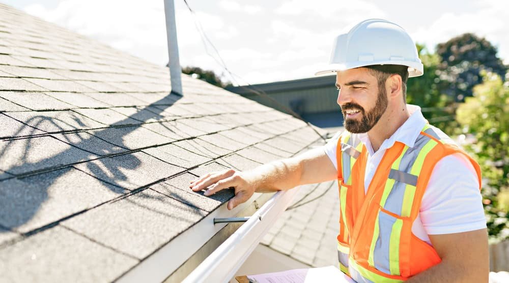 A construction worker wearing a white hard hat and orange safety vest inspects a roof. He is touching a shingle while holding a clipboard. The background shows trees and part of a building under a sunny sky.