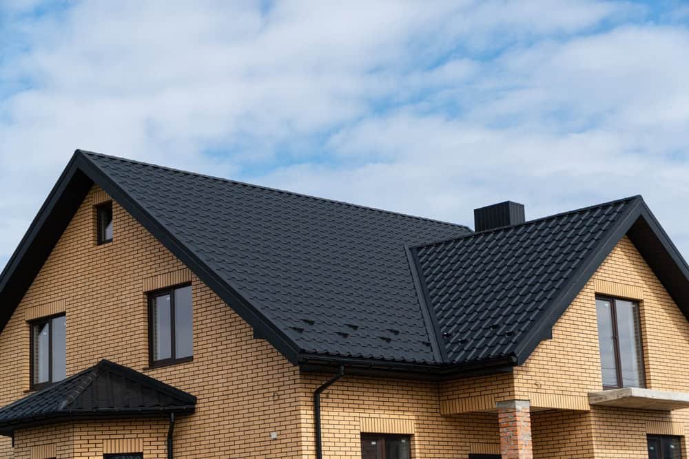 A modern two-story house with light brown brick walls and a dark gray metal roof against a cloudy blue sky. The building features large windows and a chimney.