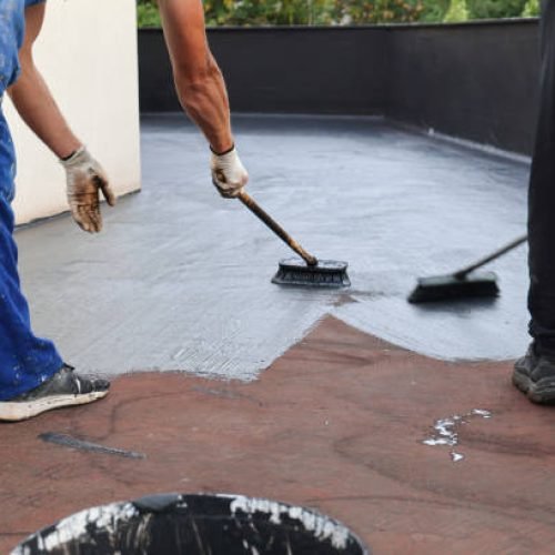 Two builders construction workers with brushes applying waterproofing layer coating on a building roof