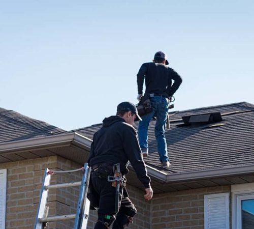 Two workers in safety gear are on the roof of a brick house. One is climbing a ladder while the other is standing on the rooftop, both under a clear blue sky.