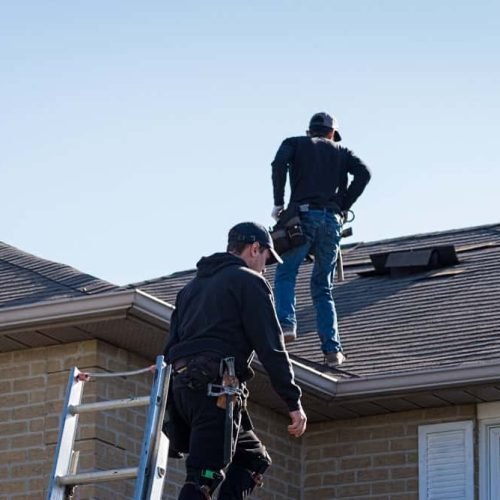 Two workers in safety gear are on the roof of a brick house. One is climbing a ladder while the other is standing on the rooftop, both under a clear blue sky.