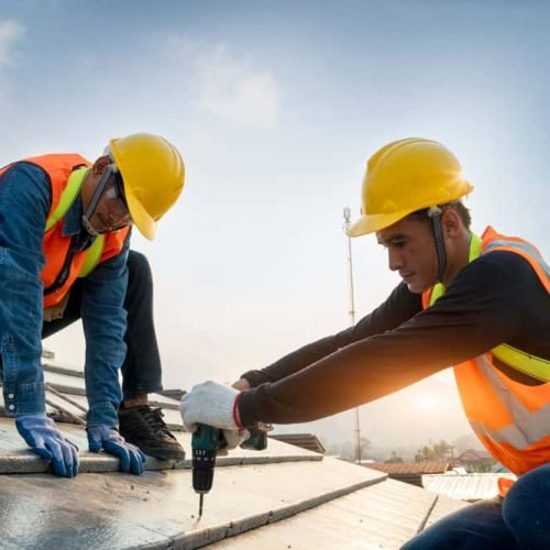 Two construction workers wearing safety vests and helmets are installing solar panels on a rooftop. One is crouched, aligning the panels, while the other uses a drill. The sky is clear, indicating a bright day.