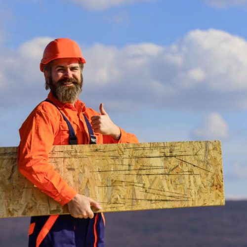 A bearded construction worker in an orange helmet and shirt gives a thumbs-up while holding a wooden plank. He stands outdoors with a cloudy sky and hills in the background.