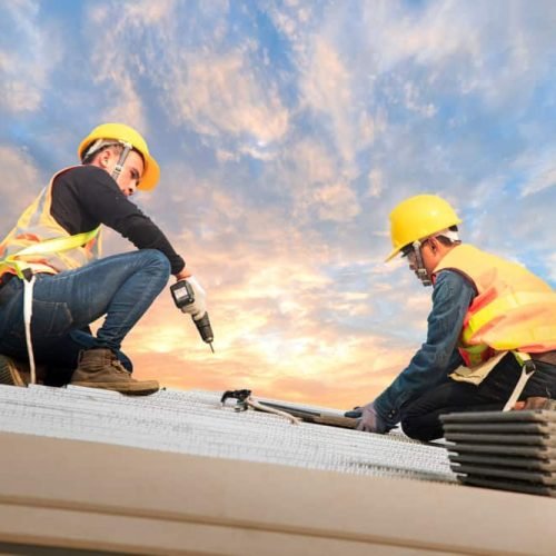 Two construction workers wearing yellow helmets and safety vests are working on a roof at sunset. One is using a power drill, while the other holds a panel. Stacks of roofing materials are nearby, and the sky is partly cloudy.