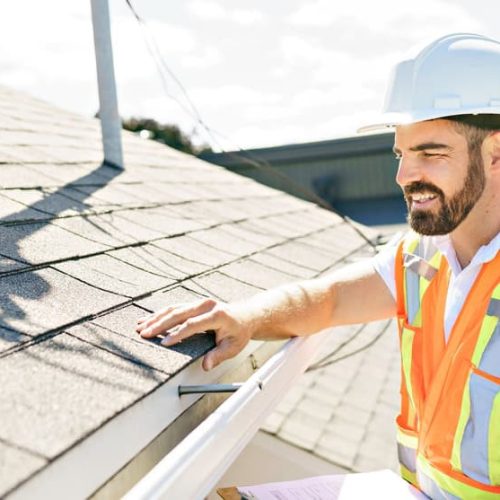A construction worker wearing a white hard hat and orange safety vest inspects a roof. He is touching a shingle while holding a clipboard. The background shows trees and part of a building under a sunny sky.