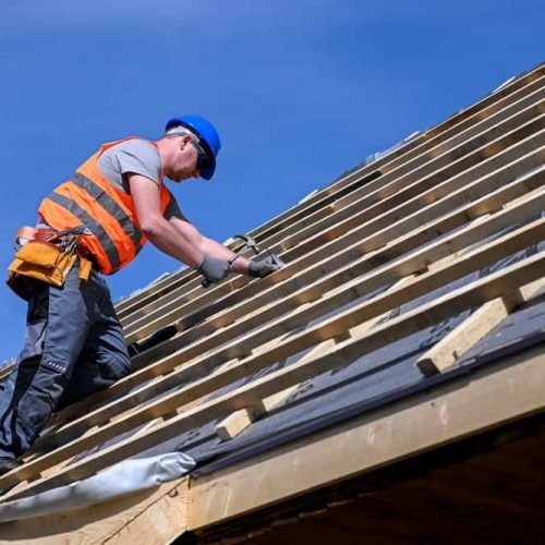 A construction worker wearing an orange safety vest and blue hard hat works on a wooden roof framework under a clear blue sky. He is kneeling, using a tool to secure pieces of wood in place.